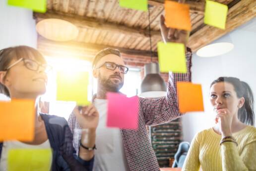 Leaders ask Questions - Three professionals brainstorming with post-its on a wall, focusing on a business plan, with a male leader flanked by two female colleagues.